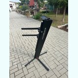 Černý kovový stojan na klávesové nástroje s dvěma vodorovnými rameny. Na hlavní nosné tyči je bílý nápis ULTIMATE SUPPORT™. Stojan stojí na dlážkovém…