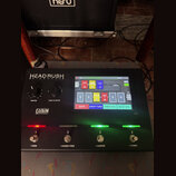 Černý MIDI kontrolér s nápisem HEAD RUSH GIGBOARD. Zařízení má barevný displej zobrazující rozhraní s nápisy DIGITAL RIG, GATE, GREEN RIC-DOO, DROP T…