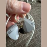 Dvě šedé sluchátka s hranatým, geometrickým tvarem a průhledným opleteným kabelem. Na obou sluchátkách je černý nápis KATO® MOONDROP PRESENTS. Povrch…