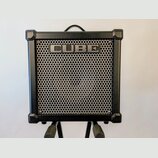 Černý kytarový kombozesilovač s kovovou mřížkou reproduktoru. V horní části mřížky je umístěn nápis CUBE. Nástroj má v rozích ochranné plastové rohy…