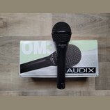 Černý dynamický mikrofon s mřížkou, na těle kterého je bílý nápis AUDIX OM3. Mikrofon leží na originální krabici, na které jsou vidět nápisy OM3 a AU…