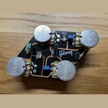 Černá tiskovaná spoje s четыřmi stříbrnými kovovými potenciometry, na kterých je vyražen nápis Gibson. Na desce jsou bílé nápisy BRIDGE, NECK, SWITCH…