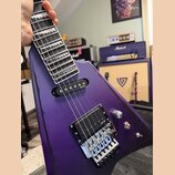 Fialová elektrická kytara s hranatým tělem a hrazdicími pražci. Na obou snímačích je bílý nápis EMG a na kobylce je viditelný nápis Floyd Rose. Povrc…