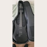 Černá elektrická kytara s matným povrchem v černém softcasu. Nástroj má dva humbuckery, fixační most a na hlavě není vidět žádné logo ani nápis. Povr…