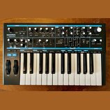 Černý analogový syntetizátor s klaviaturou a ovládacím panelem. Na levé straně je nápis novation BASS STATION II Analog Synthesizer. Panel obsahuje m…