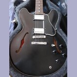 Černá polodupatá elektrická kytara s černým pickguardem a dvěma humbuckery. Nástroj je umístěn v otevřeném kufru s šedým polstrováním. Na těle jsou č…