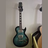Elektrická kytara s tmavě zeleným gradientem a viditelnou strukturou dřeva. Na hlavě je bílý nápis Cort. Nástroj má dva humbuckery, čtyři otočné knof…