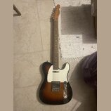 Elektrická kytara typu Telecaster s tónem sunburst (přechod od černé k hnědé a žluté). Má bílý pickguard, dva snímače a na hlavě hmatníku je viditeln…