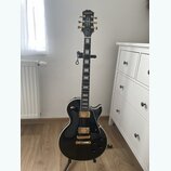 Černá elektrická kytara s gold hardwarem a bílým bindingem, umístěná na stojanu. Na hlavě hmatníku je bílý nápis Epiphone. Kytara má dva humbuckery,…