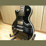 Černá elektrická kytara s humbuckery a bílým lemováním kolem korpusu. Na kytaru jsou vidět čtyři ovládací knofíky s nápisy „VOLUME“ a „TONE“. Nástroj…