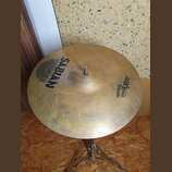 Zlatý činel typu crash umístěný na kovovém stojanu. Na povrchu jsou čitelné nápisy SABIAN a AAX STAGE CRASH 16"/40cm. Povrch vykazuje drobné nečistot…