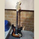 Elektrická kytara s trupem v barevnosti sunburst a černým pickguardem. Na hlavě hmatníku je vidět nápis Squier by Fender. Nástroj má tři snímače typu…