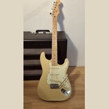 Krémová elektrická kytara typu Stratocaster s bílým pickguardem a javorovým hmatníkem. Na hlavě je viditelné logo Squier. Kytara je umístěna před čer…