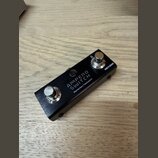 Černý kovový externí spínáč s dvěma stříbrnými tlačítky. Na horní straně je bílý nápis AMPERO SWITCH a pod ním menší text Momentary Footswitch. Na pr…