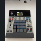 Šedý sampler MPC SAMPLE značky AKAI professional. Zařízení má v horní části displej s vizualizací zvukové vlny, ovládací knoflíky a v pravé části 16…