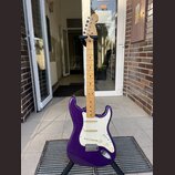 Fialová elektrická kytara s bílým pickguardem a javorovým hmatníkem. Na hlavě je viditelné logo Fender. Nástroj je umístěn na černém stojanu.