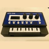 Černý kompaktní syntetizátor s modrým horním panelem a malou klaviaturou. Na horní části je bílý nápis NTS ONE a text PROGRAMMABLE SYNTHESIZER. Panel…