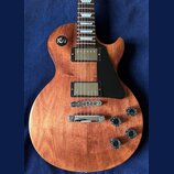 Elektrická kytara s hnědým dřevěným povrchem a černým plechtrátem. Nástroj má dva humbuckery s kovovými kryty, čtyři černé otočné knoflíky a přepínač…