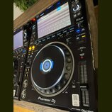 Černý digitální DJ player s displejem a velkým ovládacím jog-kolem. Na přístroji jsou viditelné nápisy Pioneer DJ a MULTI PLAYER CDJ-3000. Povrch je…