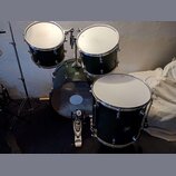 Sada tmavě zelených bubnů s kovovým kováním. Na basovém bubnu a tomu je vidět logo Pearl. Povrchy blan jsou bílé, na levém tomu jsou vidět drobné sto…