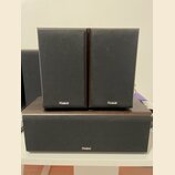 Tři reproduktory značky Magnat s černými textilními kryty a tmavým dřevěným povrchem. Dva menší stolní reproduktory stojí vedle sebe nad jedním širší…