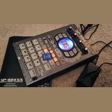 Šedý sampler s názvem SP-404SX LINEAR WAVE SAMPLER. Zařízení obsahuje mřížku 12 padů, ovládací knofíky a kruhový displej s červeným textem. Na přední…