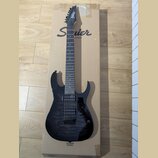 Černá elektrická kytara s dvojsekmovou hmatníkem a dvěma humbuckery, položená na kartonovém obalu. Na hlavě kytary je bílé logo Ibanez a na kartonu n…