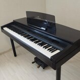 Černé digitální piano s klaviatura v klasickém provedení. Na hudebním stojanu je bílý nápis Clavinova a nad ním menší logo Yamaha. Nástroj je umístěn…