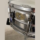 Detailní pohled na kovový snare drum stříbrné barvy s chromovaným obručováním. Na korpusu je oválný zlatý štítek s nápisy Pearl, Custom Alloy, Steel…