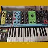 Semi-modulární analogový syntetizátor s klaviaturou a ovládacím panelem v různých barvách (šedá, modrá, zelená). Na spodní liště je nápis GRANDMOTHER…