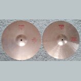 Dva mosazné činely typu hi-hat položené vedle sebe na bílém koberci. Oba mají červené nápisy a logo Paiste 2002. Levý činel má nápis TOP MEDIUM HI-HA…