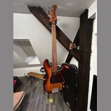 Elektrická basová kytara s trupem v červeně-žlutém sunburst provedení a tmavě červeným pickguardem. Na hlavě hmatníku není žádný viditelný nápis. V p…