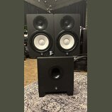 Sada tří černých studiových reproduktorů, dvě menší bedny umístěné nad jednou větší. Na spodní části každého reproduktoru je bílé logo Yamaha v kruhu…