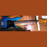 Modrá elektrická basová kytara s černým pickguardem a hnědým hmatníkem. Na hlavě nástroje je viditelné logo Fender a nápis PRECISION BASS. Nástroj vy…