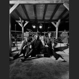 Černobílá fotografie zachycuje čtyři osoby sedící pod dřevěným přístřeškem. V pozadí je vidět bicí souprava a kytarový zesilovač s nápisem Highgain &…