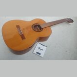 Kytara s přírodním dřevěným povrchem a hnědým hmatníkem. Vedle nástroje leží krabička s nápisy Blond STRINGS, classical guitar Medium a nečitelný tex…