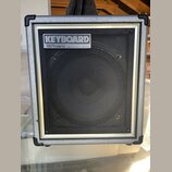 Kombinovaný zesilovač s šedým povrchem a černou síťovkou. V horní části je umístěn štítek s nápisy KEYBOARD a Roland SPECIALIZED MUSIC AMPLIFIER. Na…