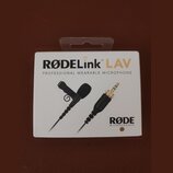 Fotografie znázorňuje bílé originální balení mikrofonu. Na krabici je uveden nápis RØDELink LAV a text PROFESSIONAL WEARABLE MICROPHONE, v pravém dol…