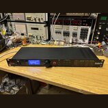 Černý rackový procesor s nápisem dbx PROFESSIONAL PRODUCTS a DriveRack PA2 SYSTEM MANAGEMENT SYSTEM. Zařízení má modrý podsvícený displej, ovládací k…