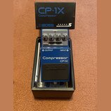 Modrý efekt pedál značky BOSS v originální krabici. Na pedálu jsou nápisy Compressor, CP-1X a čtyři ovládací prvky s popisky LEVEL, ATTACK, RATIO a C…
