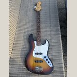 Čtyřstrunná elektrická basová kytara s tělem v provedení sunburst a bílým pickguardem. Na hlavě hředu je viditelné logo Fender. Nástroj vykazuje znám…