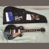 Černá elektrická kytara s hnědým pickguardem a dvěma humbuckery. Na hlavě nástroje je bílé logo Gibson. Vedle kytary je černý obal s bílým nápisem Gi…