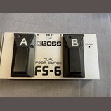 Stříbrný dvoupedálový spínací jednotka s černými plasty. Na přístroji jsou nápisy BOSS, DUAL FOOT SWITCH a FS-6 oraz označení pedálů A a B. V dolní č…
