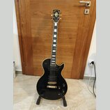 Černá elektrická kytara s gold hardwarem, umístěná na černém stojanu. Na hlavě je zlatý nápis Gibson. Nástroj má dva humbuckery, dva ovládací knofíky…