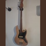 Elektrická basová kytara s přírodním dřevěným povrchem těla a hředu. Nástroj má černý pickguard, dva snímače a čtyři ladicí kolíky na hlavě. Na těle…