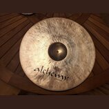 Kovový činel zlatavé barvy s viditelným rakováním povrchu. Na spodní části je černý nápis alchemy cymbals®. V horní části jsou drobné, lehce čitelné…