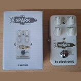 Bílá efekt pedál s nápisy SPARK BOOSTER a tc electronic. Pedál má čtyři bílé ovládací knoflíky s popisky GAIN, LEVEL, BASS a TREBLE, dále přepínač FA…