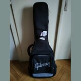 Černý softcase na kytaru s bílým nápisem Gibson v dolní části. Na popruhové části je připevněna bílá papírová štítková visačka s nečitelným textem. P…
