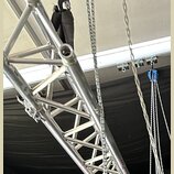 Detail stříbrného hliníkového systémového trussu s mřížkovou konstrukcí. Vedle konstrukce jsou vidět kovové řetězy a modrý zvedací mechanismus. Na po…