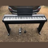 Černý digitální piano s klaviaturou a přiloženým notovým pultem na černém stojanu. Na pravé straně horního panelu je nápis CASIO PRIVIA. Pod nástroje…